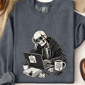 May include: A dark heather gray crewneck sweatshirt featuring a graphic of a skeleton wearing a hoodie and glasses, sitting at a laptop with a sticky note that says STILL WORKING. Next to the laptop is a mug labeled FUELED BY COFFEE & DEADLINES.