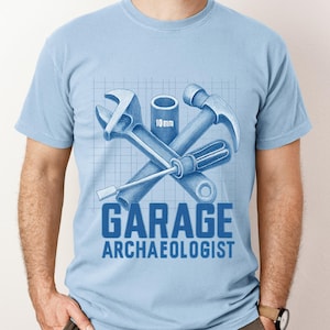 May include: Pale blue t-shirt featuring a blue graphic of crossed tools, including a wrench, screwdriver, hammer, and socket. The text "GARAGE ARCHAEOLOGIST" is printed below the tools in blue.