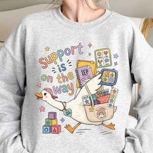 Könnte beinhalten: Ein hellgraues Sweatshirt mit Rundhalsausschnitt und einer bunten Grafik einer Gans, die eine rosa Kappe und ein herzförmiges Schild mit der Aufschrift Silly Goose trägt. Der Text Support is on the way steht über der Gans mit Schultasche.