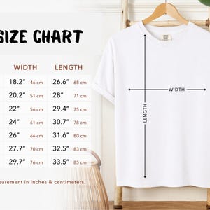 Size Chart. White t-shirt on a wooden hanger with a size chart. The chart displays measurements in inches and centimeters for sizes S to 4XL. The width measurements range from 18.2" (46 cm) to 29.7" (76 cm), and the length measurements range from 26.6" (68 cm) to 33.5" (85 cm).