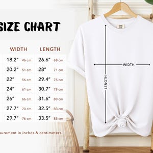 Size Chart. White t-shirt on a wooden hanger with a size chart. The chart displays measurements in inches and centimeters for sizes S to 4XL. The width measurements range from 18.2" (46 cm) to 29.7" (76 cm), and the length measurements range from 26.6" (68 cm) to 33.5" (85 cm).