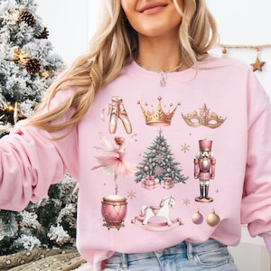 May include: A light pink sweatshirt featuring a collection of watercolor illustrations. The design includes ballet shoes, a crown, a masquerade mask, a ballerina, a Christmas tree, a nutcracker, a drum, a rocking horse, and ornaments.