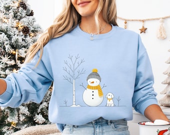 Puede incluir: Sudadera azul claro con un muñeco de nieve con gorro gris y bufanda amarilla, y un perro pequeño. El muñeco de nieve tiene ojos de botón negro y nariz de zanahoria. Un árbol desnudo está al lado del muñeco de nieve. Diseño de invierno.