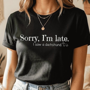 May include: Black t-shirt with the text "Sorry, I'm late. I saw a dachshund" in white. The shirt is paired with blue jeans. The model is wearing gold necklaces and bracelets.