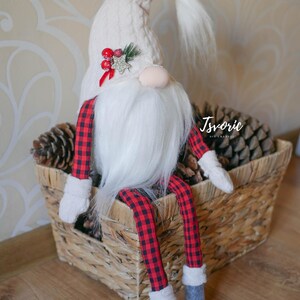 May include: A white gnome with a red and black checked outfit sits in a wicker basket filled with pine cones. The gnome has a white beard and a white hat with a red berry and gold star embellishment.