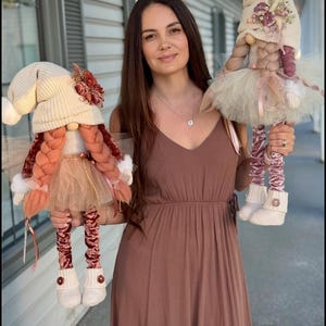 Op de afbeelding: Twee handgemaakte dwergpoppen met roze en witte hoeden, roze en witte jurken en roze en witte schoenen. De poppen worden vastgehouden door een persoon die een bruine jurk draagt.