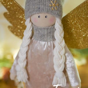 May include: A handmade angel doll with a gray knit hat featuring a gold snowflake. The doll has long white braided hair, rosy cheeks, and wears a pink velvet dress with gold trim. Gold glitter wings are visible.