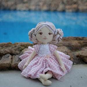 Puede incluir: Muñeca de tela hecha a mano con tez clara, ojos marrones y moños trenzados. La muñeca lleva un top de ganchillo blanco y un vestido a cuadros rosa y blanco. Una cinta rosa está atada alrededor de la cintura. La muñeca está sentada sobre una superficie de hormigón.