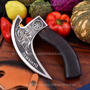 May include: A silver axe with a black wooden handle. The axe head features a skull and a circular design with runes. The axe is on a wooden surface with a leather sheath.
