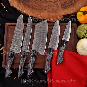 Może przedstawiać: Zestaw sześciu noży kuchennych ze stali damasceńskiej z ciemnymi drewnianymi uchwytami i mosiężnymi nitami. Ostrza mają unikalny wzór. Noże są prezentowane na drewnianej desce do krojenia z warzywami w tle. Widoczny jest tekst "MyPersonalMemories4u".