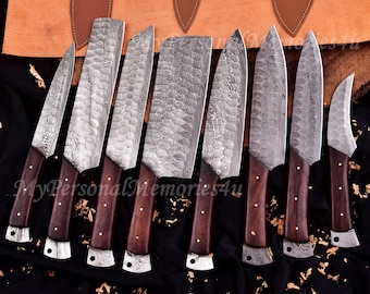 May include: A collection of eight Damascus steel kitchen knives. Each knife has a dark wood handle and silver bolster. The blades display a textured pattern. The knives are displayed in front of a leather knife roll.