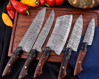May include: Five kitchen knives with wooden handles and Damascus steel blades. The knives are arranged on a wooden cutting board. The knives include a chef's knife, a cleaver, a paring knife, and two utility knives.