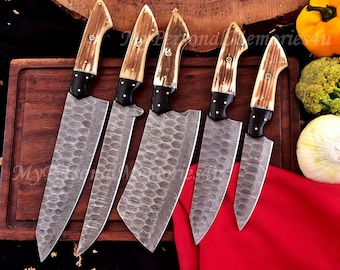 May include: A set of five kitchen knives with patterned blades and antler-style handles. The blades have a textured design, and the handles are a mix of light brown and black. The knives are arranged on a wooden cutting board.