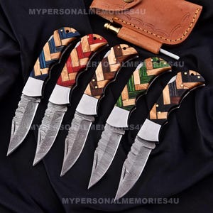 May include: Five pocket knives with patterned wooden handles. The handles are in different colours, including blue, red, green, and brown. The blades are made of Damascus steel.
