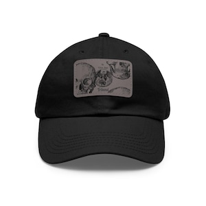 Peut inclure: Casquette de baseball noire avec une visière incurvée. La casquette présente un écusson rectangulaire avec une illustration grise de crânes et le texte "Tribunal". Le chapeau est fait d'un tissu durable et possède une sangle réglable.