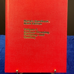 May include: A red book titled "Metals Handbook, Ninth Edition, Volume 5: Surface Cleaning, Finishing, and Coating." The book is displayed on a gold-colored stand against a blue background. The text is in gold lettering.
