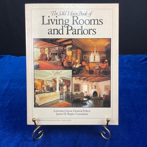 May include: A hardcover book titled "The Old House Book of Living Rooms and Parlors" with images of interior spaces. The book is displayed on a gold-colored metal stand against a blue background. The cover features a cream-colored background with text and images.
