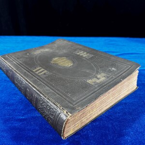 May include: A vintage, dark brown leather-bound book with gold accents and intricate designs on the cover. The book is closed and lying on a blue surface.