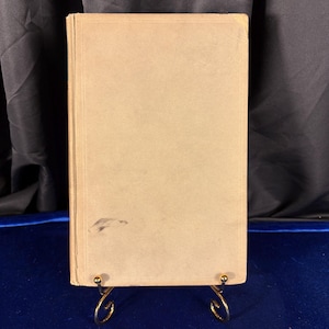 May include: A vintage book with a beige cover, displayed on a gold-colored metal stand. The book's cover shows some wear and tear, with a few dark marks. The background is a dark curtain and a blue surface.