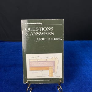 May include: A book titled "QUESTIONS & ANSWERS ABOUT BUILDING" from Fine Homebuilding. The cover is dark green with white text. A diagram of a building cross-section is visible. The book is displayed on a gold-colored metal stand against a blue background.