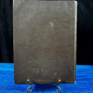May include: A brown leather-bound book with a textured cover. The book is standing on a gold metal stand.