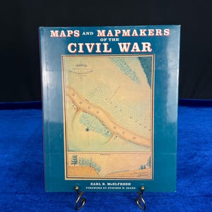 May include: A teal book titled "MAPS and MAPMAKERS of the CIVIL WAR" with a detailed map illustration. The book is displayed on a decorative metal stand. The author's name, Earl B. McElfresh, and the foreword author, Stephen W. Sears, are also visible.