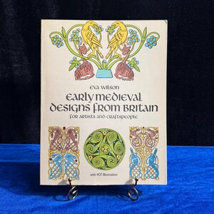 May include: A book titled "Early Medieval Designs from Britain" by Eva Wilson. The cover features a yellow and green illustration of birds and a Celtic knot design. The book is for artists and craftspeople and includes 407 illustrations.