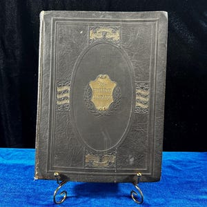May include: A dark gray leather-bound book with gold lettering and decorative accents. The title "The Volume Library" is embossed on the front cover.