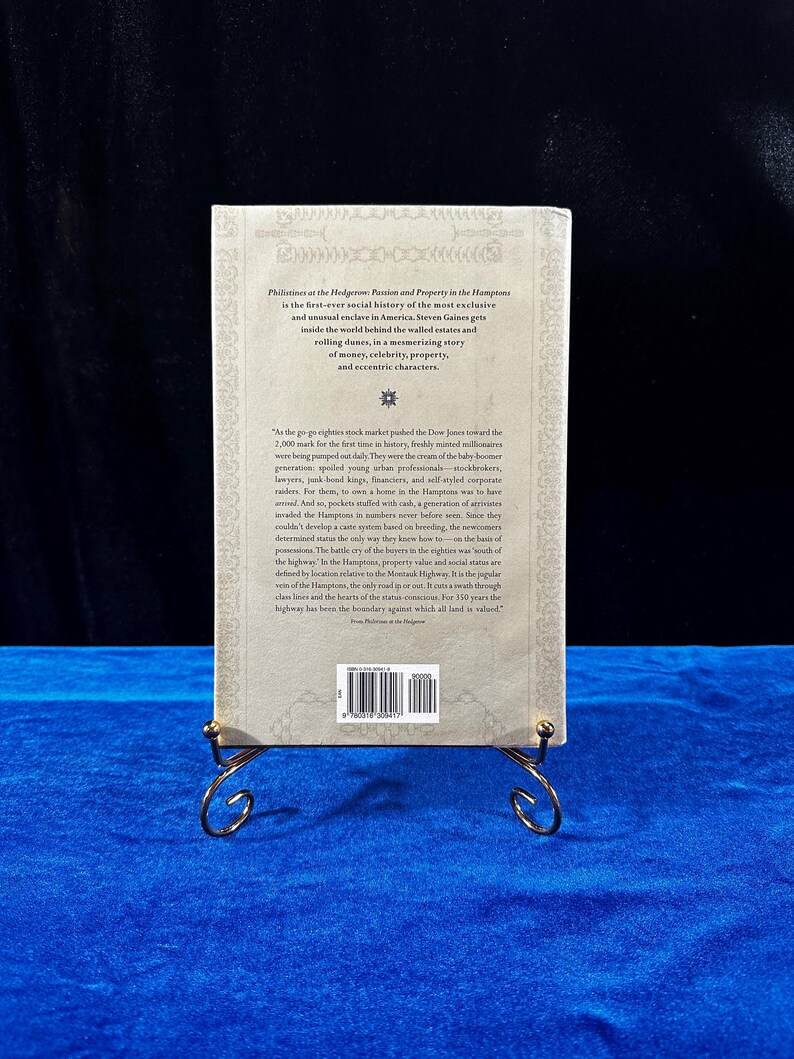 May include: A beige book cover with a black and white image of a star on the front. The book title is "Philistines at the Hedgerow: Passion and Property in the Hamptons" and the author is Steven Gaines. The back cover has a blurb about the book and a barcode.