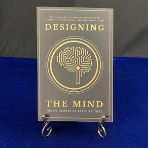 May include: A book titled "DESIGNING THE MIND" is displayed on a gold metal stand. The book cover is dark gray with gold lettering and a gold brain graphic. The text "THE PRINCIPLES OF PSYCHITECTURE" is below the title. A quote is at the top.