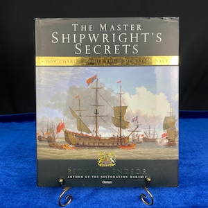 May include: A hardcover book titled "THE MASTER SHIPWRIGHT'S SECRETS" with an illustration of sailing ships. The book cover features the text "HOW CHARLES BUILT THE RESTORATION NAVY" and the author's name, Richard Endsor. The book is displayed on a gold-colored stand.