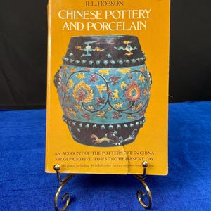 May include: A book titled "Chinese Pottery and Porcelain" by R.L. Hobson. The cover features a detailed illustration of a decorative ceramic piece with blue, gold, and black accents. The book is displayed on a gold-colored metal stand.