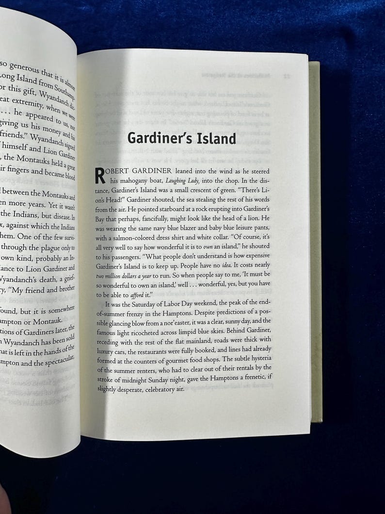 May include: A book page with a black and white photo of a boat on the water. The text on the page reads "Gardiner's Island" and "Robert Gardiner leaned into the wind as he steered his mahogany boat, Laughing Lady, into the chop."