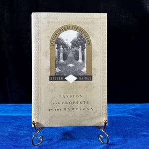 May include: A book titled "Philistines at the Hedgerow" by Steven Gaines. The book cover features a gold-colored illustration of a garden path with a white building in the background. The book's subtitle is "Passion and Property in the Hamptons".