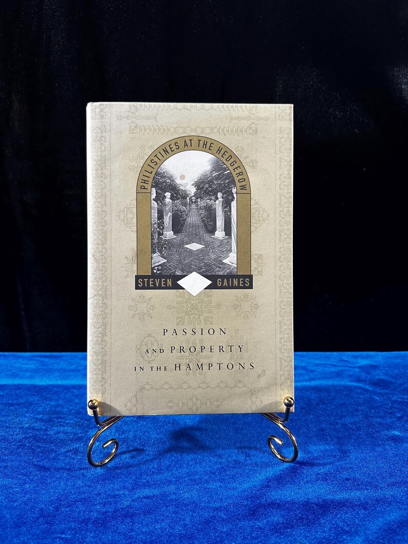 May include: A book titled "Philistines at the Hedgerow" by Steven Gaines. The book cover features a gold-colored illustration of a garden path with a white building in the background. The book's subtitle is "Passion and Property in the Hamptons".