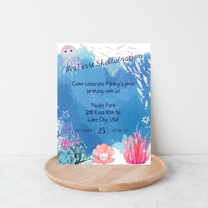 May include: A birthday invitation with the text "My First Shellabration" in a watercolour style. The design features aquatic illustrations, including a jellyfish, fish, coral, and a seashell. The invitation invites guests to celebrate Paisley's first birthday.