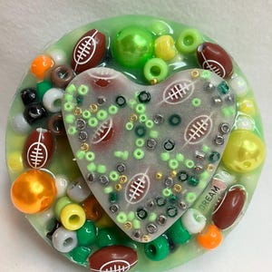 Puede incluir: Un llavero de resina verde en forma de corazón con fondo blanco y un patrón de fútbol. El llavero está rodeado de cuentas verdes, amarillas, naranjas y marrones, así como de dijes con forma de fútbol. La palabra "DREAM" es visible en el llavero.