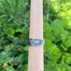 May include: A silver ring with a detailed floral and fruit design. The ring is displayed on a wooden ring sizer against a backdrop of green foliage. The ring's band features a repeating pattern of leaves and berries.