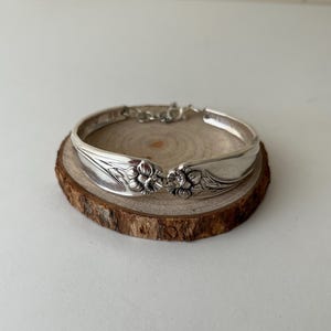 May include: A silver-toned bracelet crafted from repurposed cutlery, featuring two mirrored floral motifs at the centre. The band is formed by the curved handles of spoons, showcasing intricate embossed petal details and a polished metallic finish.