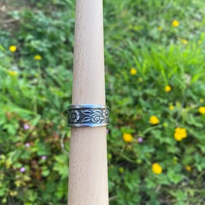 Puede incluir: Un anillo plateado con un diseño floral se muestra en un calibrador de anillos de madera. El anillo presenta tallas detalladas de flores y hojas. El fondo es una vista borrosa de hierba verde y flores amarillas.
