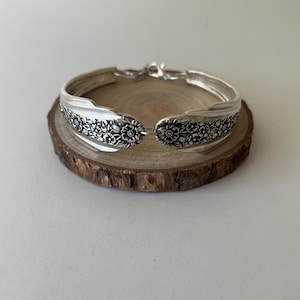May include: A silver-toned bracelet crafted from a repurposed spoon, featuring an intricate embossed floral pattern across the curved metal surface. The piece includes a metallic clasp mechanism at the back to secure the ends of the spoon handle.