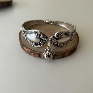 May include: A silver-toned bracelet crafted from a repurposed spoon, featuring an ornate embossed floral pattern along the band and a small, detailed ladybug charm dangling from the centre. The metal has a polished, reflective finish.