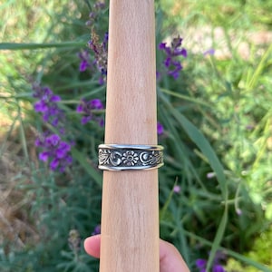 May include: A silver-toned metal ring featuring an embossed floral and scrollwork pattern. The band has a flat profile with a darkened finish in the recessed areas of the design, highlighting the central flower motif and surrounding leafy vines.