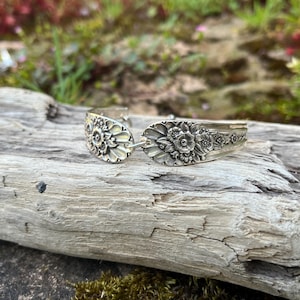 May include: A silver-toned metal cuff bracelet crafted from repurposed cutlery, featuring an intricate embossed floral pattern across the surface. The design displays detailed petals and leaves in a relief style, with a polished finish that highlights the metallic texture and the curved shape of the original spoon handle.