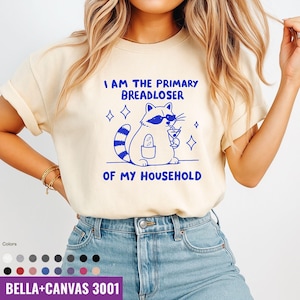 May include: Cream-colored t-shirt with a blue graphic of a raccoon wearing sunglasses and holding a cocktail. The text "I AM THE PRIMARY BREADLOSER OF MY HOUSEHOLD" is above and below the raccoon illustration. The shirt is a Bella+Canvas 3001.
