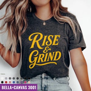 May include: Dark gray Bella+Canvas 3001 t-shirt with the words "Rise & Grind" in yellow script. The shirt is a v-neck style and is being worn by a person. A color chart is visible at the bottom.