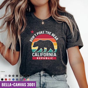May include: Dark gray t-shirt with a graphic design featuring a bear silhouette inside a colorful sunset circle. The text reads "DON'T POKE THE BEAR" and "CALIFORNIA REPUBLIC". The shirt is a Bella+Canvas 3001.