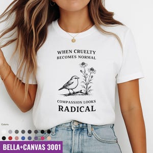 Puede incluir: Camiseta blanca con un gráfico en blanco y negro de un pájaro posado en una rama junto a flores. El texto dice: "CUANDO LA CRUELDAD SE VUELVE NORMAL, LA COMPASIÓN PARECE RADICAL". La camiseta es una Bella+Canvas 3001.