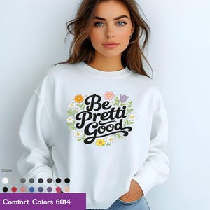 May include: White crewneck sweatshirt with the phrase "Be Pretti Good" in black script, surrounded by a floral wreath of daisies, orange, pink, and purple flowers. The sweatshirt is a Comfort Colors 6014. A palette of colors is shown at the bottom.