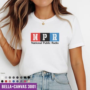 Puede incluir: Camiseta blanca con las letras "NPR" en bloques rojos, negros y azules, con "National Public Radio" impreso debajo. La camiseta está hecha de un material suave y es una Bella+Canvas 3001.